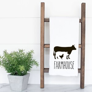 以下が含まれることがあります： 黒色の牛と鶏のシルエットが描かれた白いキッチンタオル。下に「FARMHOUSE」という文字が印刷されています。