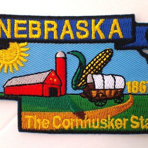 May include: Embroidered patch depicting the state of Nebraska. The patch features a blue banner with the word "NEBRASKA" in yellow lettering, a red barn, a yellow sun, a wagon, a stalk of corn, and the text "The Cornhusker State" in yellow lettering. The year "1867" is also included in yellow lettering.