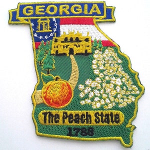 May include: An embroidered patch shaped like the state of Georgia. The patch is green with a yellow outline and features a peach, a white flower, a building, and a small American flag. The text "The Peach State" and "1788" are also embroidered on the patch.