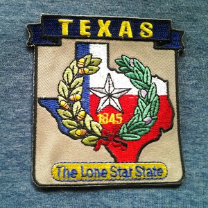 May include: A beige embroidered patch featuring a red, white and blue outline of the state of Texas. The patch includes a five-pointed star, a wreath of green leaves, and the text "Texas" in yellow letters on a blue banner. The text "The Lone Star State" is also embroidered on the patch in yellow letters on a blue banner.