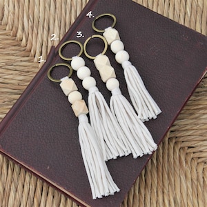 May include: Four keychains with wooden beads and white tassels. The beads are geometric shapes and the tassels are made of cotton yarn.