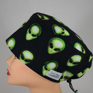 May include: Black surgical scrub cap with a repeating pattern of green alien faces. The alien faces have large black eyes and are outlined in red. The cap has a white tag with the text "Erika Moment" and a black drawstring.