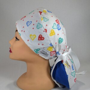 May include: White scrub cap with a colorful heart pattern in red, yellow, blue, and teal. The cap has a blue lining and a white ribbon tie. The cap is designed for medical professionals.
