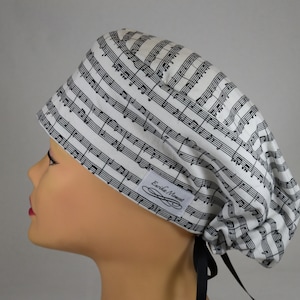 May include: A white and black musical note patterned scrub cap. The cap is tied in the back with black ribbons. A small white tag with the text "Erika Moment" is sewn on the side of the cap.
