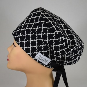 May include: A black and white patterned scrub cap with a quatrefoil design. The cap has a black tie at the back and a small white label with the text "Erika Moment". The cap is worn on a mannequin head.