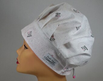 May include: A white scrub cap with a silver glitter finish and a repeating pattern of the text "LOVE YOU more" with a small pink heart. The cap features a wide band, pleated fabric, and a small white label reading "Eunike Moment".
