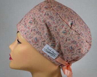 May include: A scrub cap featuring a dusty pink base with a multi-colored paisley and floral pattern in shades of teal, red, and cream. The cap includes a white rectangular label with the text Eurike Moment and a light peach ribbon tie at the back.