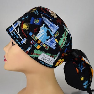 May include: A black scrub cap featuring a New York City theme. The cap is adorned with iconic landmarks like the Statue of Liberty, the Brooklyn Bridge, and the New York City skyline. The cap has a ponytail holder.