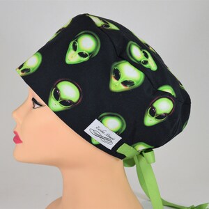 May include: Black surgical scrub cap featuring a repeating pattern of green alien faces. The cap has a light green ribbon tie and a small white tag with the text "Erika Monet".