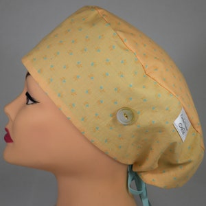 Op de afbeelding: Een gele operatiekap met een bloemenpatroon in lichtblauw en oranje. De kap heeft een witte knoop en een label met de tekst "Erika Marie". Het is vastgebonden met een lichtblauw lint.