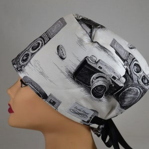 Può includere: Un cappello chirurgico bianco con un motivo in bianco e nero di illustrazioni di macchine fotografiche vintage. Il cappello ha una chiusura con lacci neri e una piccola etichetta con la scritta "Eureka Moment". Il design è un motivo ripetuto di macchine fotografiche.