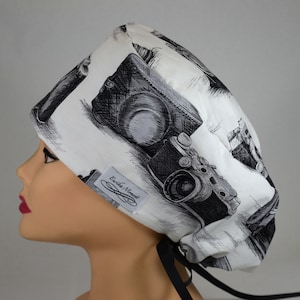 Può includere: Un cappello chirurgico bianco con una stampa di fotocamere in bianco e nero. Il cappello ha una chiusura con lacci neri e una piccola etichetta con la scritta "Erika Moment".