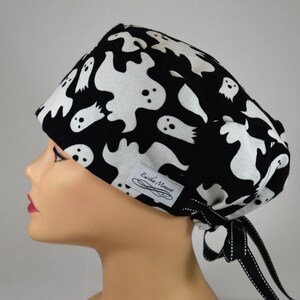 May include: A black scrub cap featuring a repeating pattern of white ghosts. The cap includes a white rectangular fabric label with the text Eureka Moment and a black ribbon tie with white stitching detail at the back for an adjustable fit.