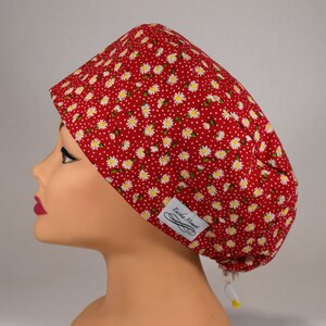 Peut inclure: Un bonnet de chirurgien rouge orné d'un motif de marguerites blanches et d'une étiquette blanche avec le texte "Erika Moment". Le bonnet est conçu pour couvrir la tête et possède un cordon de serrage pour l'ajuster. Le bonnet est en tissu floral.