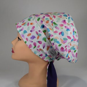 Puede incluir: Un gorro quirúrgico blanco con un colorido estampado de hojas de acuarela en tonos rosa, morado, azul y naranja. El gorro tiene un forro y lazos azul marino. La etiqueta dice "Erika Nina".