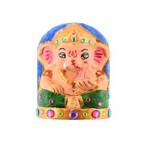 May include: A hand-painted figurine of the deity Ganesha with a peach-colored complexion, wearing green pants and a gold crown. The figure is set against a blue arched background and is decorated with small, colorful embedded rhinestones.