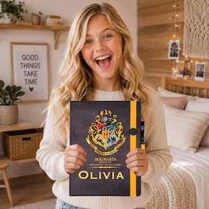 Puede incluir: Un cuaderno negro con el escudo de la Escuela Hogwarts de Magia y Hechicería en dorado, rojo, verde y azul. La portada incluye el texto Hogwarts School of Witchcraft and Wizardry y el nombre Olivia en dorado, con una banda elástica amarilla.