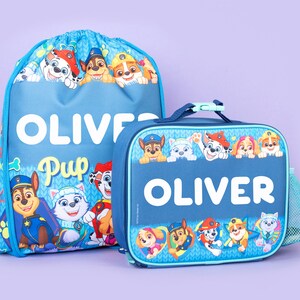 May include: A blue drawstring backpack and a matching rectangular lunch bag featuring Paw Patrol characters. Both items are personalized with the name OLIVER in white text. The designs include various cartoon dogs in colorful outfits.