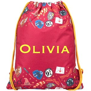 Pode incluir: Uma bolsa vermelha com cordão amarelo e o nome "OLIVIA" em letras amarelas. A bolsa é estampada com vários gráficos temáticos de Harry Potter, incluindo o brasão de Hogwarts, um pomo de ouro e uma varinha.