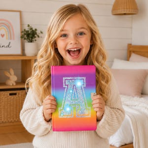 Könnte beinhalten: Ein rechteckiges Notizbuch mit einem leuchtenden Regenbogen-Farbverlauf auf dem Einband, das in der Mitte einen großen Buchstaben A aus silbernem Glitzer zeigt. Das Notizbuch hat einen kleinen Metallverschluss an der rechten Seite und eine glatte, bunte Oberfläche, die von Pink zu Violett, Blau, Grün und Orange übergeht.