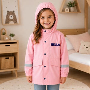 May include: A light pink hooded raincoat featuring dark blue snap buttons, two front pockets, and reflective silver bands on the sleeves. The name BELLA is printed in dark blue capital letters on the upper left chest of the jacket.