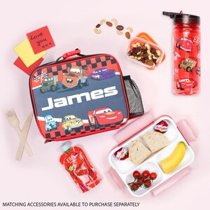 Pode incluir: Um conjunto de almo&ccedil;o infantil com uma lancheira xadrez vermelha e preta com desenhos de carros de desenho animado e o nome "James". Inclui uma garrafa de &aacute;gua vermelha, uma lancheira com comida e uma embalagem de pur&ecirc; de frutas.