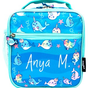 May include: A turquoise blue lunch bag with a zipper closure and a mesh side pocket. The bag features a whimsical design of narwhals with rainbow horns and a blue ocean background. The bag is personalized with the name "Anya M."