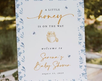 May include: A white sign with blue floral borders and gold lettering announces a baby shower. The text reads "A little honey is on the way. Welcome to Serena's Baby Shower, August 15, 2029".
