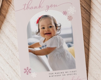 Puede incluir: Una tarjeta de agradecimiento rosa con una foto de una niña pequeña con un vestido blanco. La tarjeta tiene un arco rosa con copos de nieve y el texto "gracias" y "por hacer mi primer cumpleaños tan especial con amor charlie".