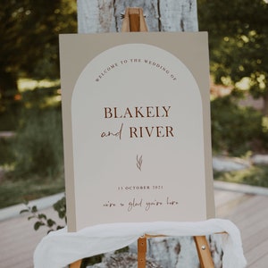 May include: A wedding welcome sign with a beige background and brown text. The sign reads "Welcome to the wedding of Blakely and River, 15 October 2021, we're so glad you're here".