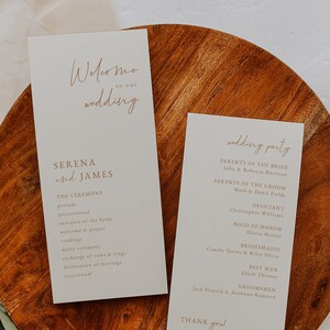 Peut inclure: Deux programmes de cérémonie de mariage ivoire avec calligraphie dorée. Le programme de gauche liste l'ordre de la cérémonie. Le programme de droite liste les membres de la fête de mariage. Les programmes sont sur une surface en bois avec des feuilles vertes.