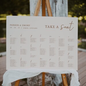 Puede incluir: Un caballete de madera muestra un plano de asientos para la boda de Parker & Emory en Aspen, Colorado. El letrero presenta nombres ordenados alfabéticamente, con las palabras "TAKE A Seat" en escritura elegante. Una tela blanca cubre la base del letrero.