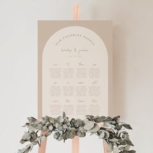 May include: A beige wedding seating chart with a brown arch at the top. The chart lists the names of guests and their assigned tables. The text at the top reads "Our Favorite People" and "Emily & Jackson 10.30.21".