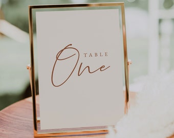 May include: A gold-framed sign with a white background and the words "Table One" written in brown cursive script.