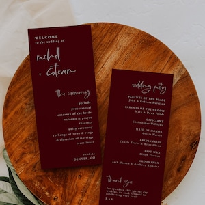 May include: Two burgundy wedding ceremony cards with white text. The first card lists the order of the ceremony, including the prelude, processional, and readings. The second card lists the wedding party, including the parents of the bride and groom, the officiant, maid of honor, bridesmaids, best man, and groomsmen. The cards also include the date and location of the wedding.