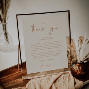 May include: A gold-framed sign with a white background and a handwritten message that reads "Thank you, A note from Jackson & Serena. It means so much to us to have you here to celebrate our marriage. Whether you have been there for us since the day we were born or you came into our lives somewhere along the way, know that you are an important part of our journey. You are our encouragers, supporters, and watchers. For the rest of our lives, this will be a night that we fondly look back on, and we are grateful that you will forever be a part of those memories. Thank you for being here and supporting us as we start our new adventure together. Enjoy the evening! With love, J & S."