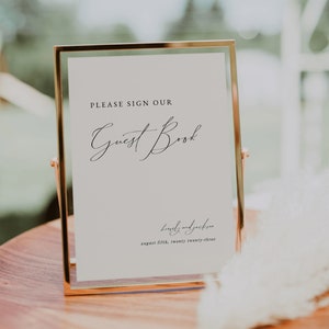 May include: A gold-framed sign with the text "PLEASE SIGN OUR Guest Book" and the date "august fifth, twenty twenty-three" written in cursive.