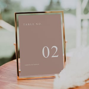 May include: A gold-framed table number sign with a light brown background. The number "02" is displayed in white text. The text "TABLE NO." and "ophelia & hudson" are also displayed in white text.