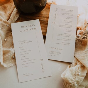 May include: A wedding ceremony programme card with the names Blakely and River, the date 05.15.21, and the location Denver, CO. The card lists the order of events for the ceremony, including the processional, entrance of the bride, welcome and prayer, readings, unity ceremony, exchange of vows and rings, declaration of marriage, and recessional. A second card lists the wedding party, including the parents of the bride and groom, the officiant, maid of honour, bridesmaids, best man, groomsmen, flower girls, and ring bearers. The cards are on a beige surface with a brown glass vase and dried flowers.