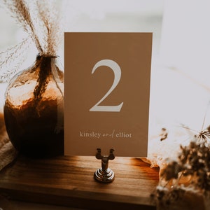 May include: A light brown card with the number "2" printed in white. The card also has the text "kinsley and elliot" printed in white. The card is sitting on a wooden table with a silver stand.