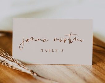 May include: A light pink table card with the name "Jenna Martino" and the text "Table 3" written in brown cursive script.