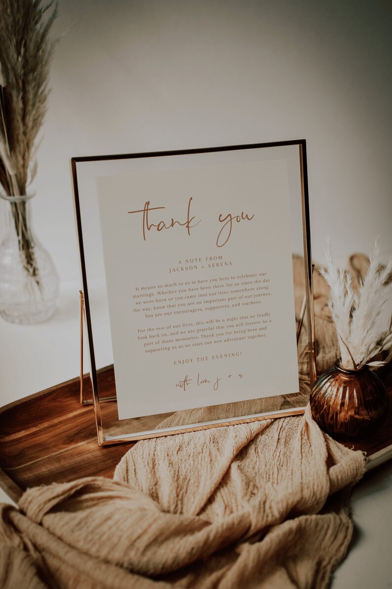 May include: A gold-framed sign with a white background and a handwritten message that reads "Thank you, A note from Jackson & Serena. It means so much to us to have you here to celebrate our marriage. Whether you have been there for us since the day we were born or you came into our lives somewhere along the way, know that you are an important part of our journey. You are our encouragers, supporters, and watchers. For the rest of our lives, this will be a night that we fondly look back on, and we are grateful that you will forever be a part of those memories. Thank you for being here and supporting us as we start our new adventure together. Enjoy the evening! With love, J & S."