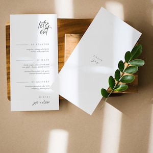 May include: A white card with the text "let's eat" in black calligraphy. The card lists a three-course menu: arugula citrus salad, fresh caught salmon with wild rice pilaf, and flourless chocolate cake with raspberry mascarpone. The date "08.07.21" and the names "jesie + diver" are printed at the bottom of the card.