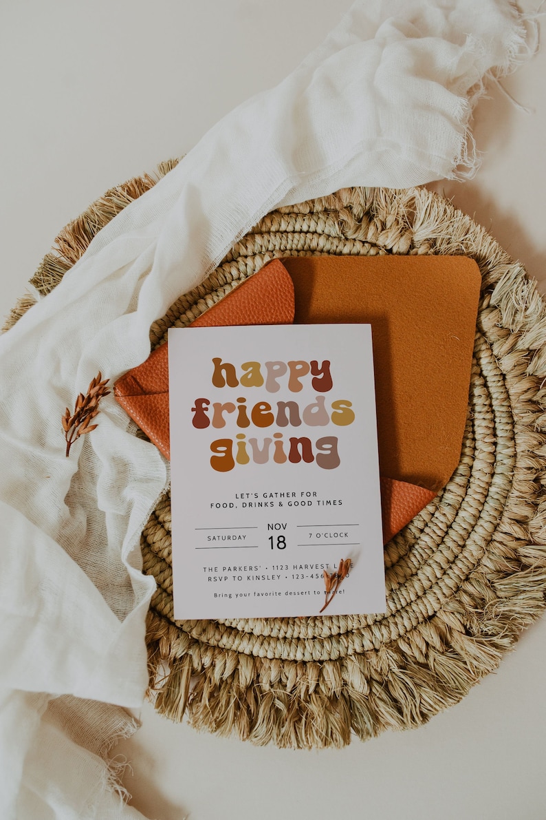 May include: A white card with the text "happy friends giving" in colorful, rounded letters. The card is on a woven basket with a brown leather pad and a white cloth. The text on the card reads "Let's gather for food, drinks & good times. Saturday, Nov 18, 7 o'clock. The Parkers, 1123 Harvest Lane, RSVP to Kinsley 123-456-7890. Bring your favorite dessert to share!"