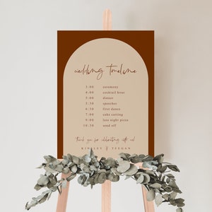 May include: A brown and beige wedding timeline sign with a rounded arch at the top. The sign lists the wedding events with times, including the ceremony, cocktail hour, dinner, speeches, first dance, cake cutting, late night pizza, and send off. The sign is on a wooden easel with greenery.