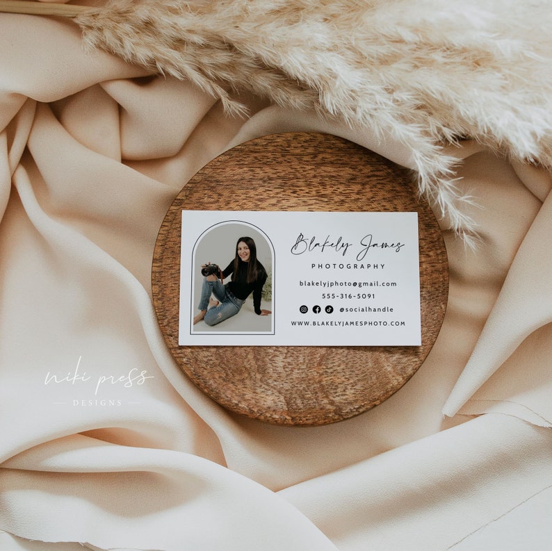 May include: White business card with a black border and rounded corners. The card features a photo of a woman holding a camera in a rounded archway. The text on the card reads "Blakely James Photography", "blakelyjphoto@gmail.com", "555-316-5091", "@socialhandle", and "www.blakelyjamesphoto.com".