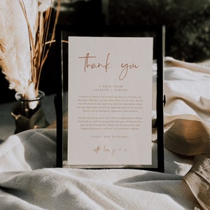 May include: A white card with a black frame, featuring the text "Thank you" in a handwritten script font. The card also includes the text "A note from Jackson + Serena" and a message expressing gratitude to guests for attending their wedding. The message includes the phrase "Enjoy the evening!" and is signed "with love, J + S".