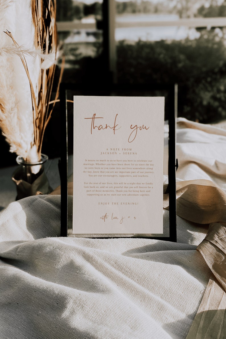 May include: A white card with a black frame, featuring the text "Thank you" in a handwritten script font. The card also includes the text "A note from Jackson + Serena" and a message expressing gratitude to guests for attending their wedding. The message includes the phrase "Enjoy the evening!" and is signed "with love, J + S".