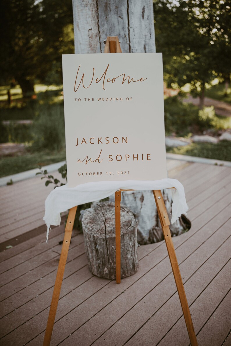 May include: Welcome to the wedding of Jackson and Sophie, October 15, 2021. A white sign with brown text on a wooden easel.