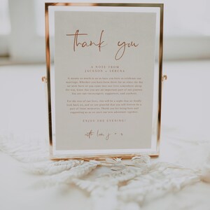 May include: A gold-framed sign with a white background and the text "Thank you" in a cursive font. The sign also includes the text "A Note From Jackson & Serena" and a message expressing gratitude to guests for attending their wedding. The sign is sitting on a white lace surface.